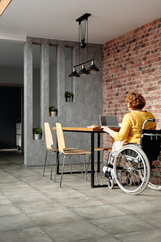 Reformas y adaptaciones para personas con discapacidad o movilidad reducida 2 Apartamento adaptado para personas en silla de ruedas, con una persona sentada en una silla de ruedas manejando un ordenador en una mesa. La pared es de ladrillo visto y el suelo es gris. El espacio es accesible y adecuado para personas con discapacidad física.