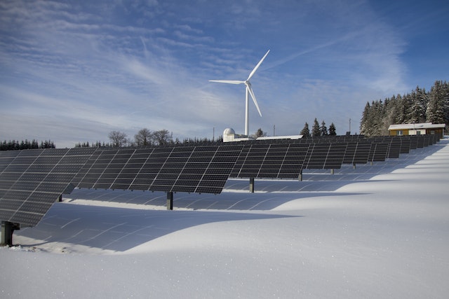 Mejoras en la eficiencia energética en obras de reforma y ampliación 2 Paneles solares en paisaje natural con cielo azul y nubes blancas, rodeados de árboles y un aerogenerador al fondo. Los paneles generan electricidad de forma sostenible y eficiente. El suelo nevado aporta un contraste invernal a la imagen.