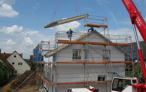 Servicios 2 Techo en construcción: dos trabajadores instalando una parte del techo de una casa blanca con ayuda de una grúa y alrededores de andamios y medidas de seguridad.