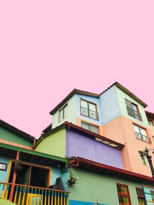 En esta imagen se muestra un encantador conjunto de casas de estilo retro, con fachadas vibrantes y una atmósfera nostálgica. Cada casa presenta una tonalidad de color única, que evoca la estética de los años 50 y añade un toque de alegría y personalidad al vecindario. El cielo rosado al atardecer crea un ambiente cálido y acogedor. Conjunto de casas de estilo retro con fachadas coloridas y un cielo rosado al atardecer