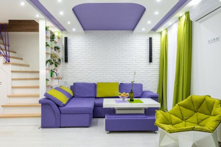 En esta imagen, podemos apreciar un salón recién reformado y pintado, donde las paredes y partes del techo están pintadas de blanco. Destacando en contraste, una sección del techo ha sido pintada en un vibrante tono morado, creando un impacto visual único. Los sofás del salón también han sido elegidos en un tono morado, complementando perfectamente con el detalle del techo. Salón recién reformado con paredes blancas y detalles en morado