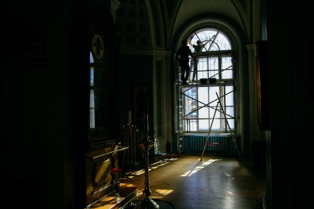 Esta imagen muestra el proceso de restauración de una vidriera en un espacio histórico. Dos operarios se encuentran subidos a un andamio trabajando en la delicada tarea de devolverle su antiguo esplendor. Los techos de bóveda y las columnas resaltan la grandiosidad arquitectónica del lugar. Restauración de una vidriera en un espacio histórico