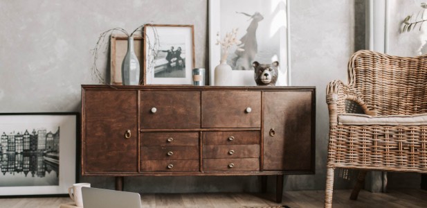 En esta imagen, podemos apreciar un mueble rústico recién pintado que irradia encanto y personalidad. El mueble se encuentra en una habitación donde se encuentra una silla de mimbre a su lado, y un cuadro de decoración colocado arriba del mueble. Mueble rústico recién pintado con detalles de decoración