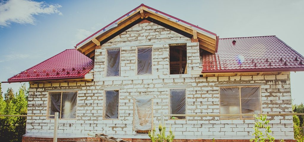 Imagen de una casa en proceso de reforma con paredes blancas desgastadas y tejado rojo rodeado de vegetación. En la imagen se observa un andamio alrededor de la casa y herramientas de construcción en el suelo. El cielo azul de fondo completa la escena