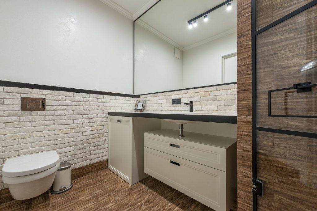 Cambio de azulejos, suelos y revestimientos 2 Imagen de un cuarto de baño con un suelo de color marrón oscuro veteado, paredes revestidas de azulejos de brillo rústico blanco en la mitad inferior y pintura blanca en la mitad superior. Un espejo grande cuelga sobre el lavabo, con muebles en tonos crema y azulejos de la ducha del mismo color del suelo.