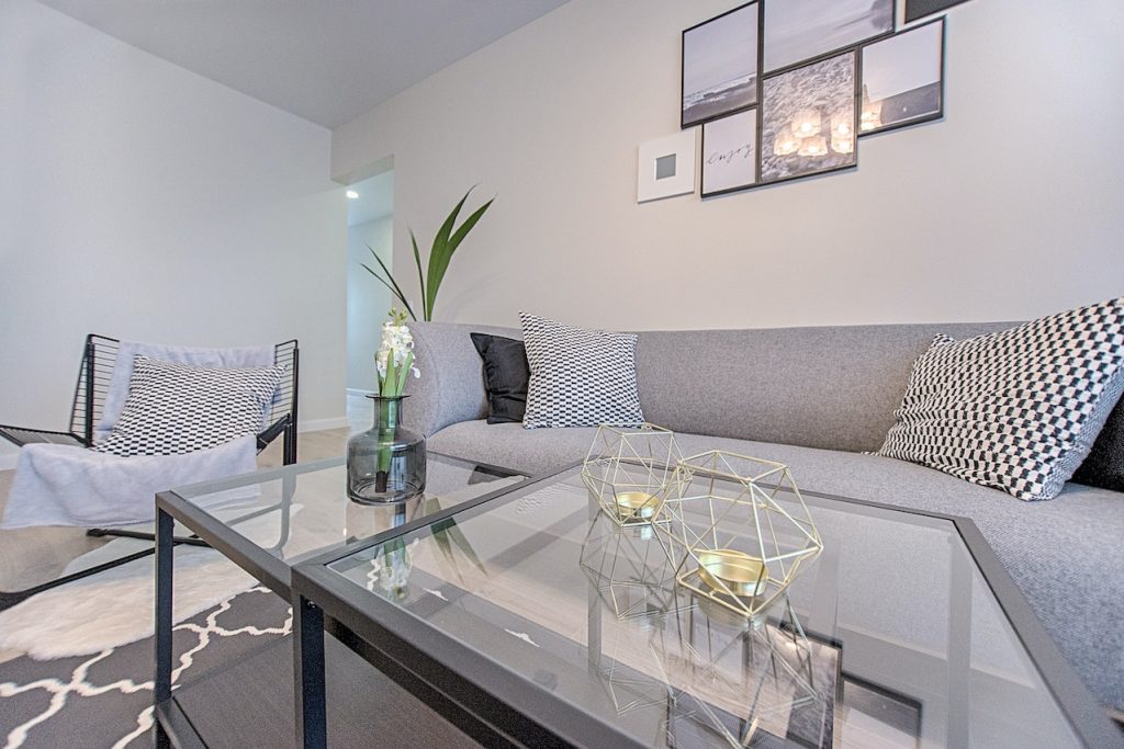 Pintura y decoración de paredes y techos 2 Imagen de un salón recién reformado con paredes y techos impecables de color blanco, un sofá de color gris, una mesa de cristal con detalles en negro y un suelo combinado de moqueta y tarima de color gris claro.
