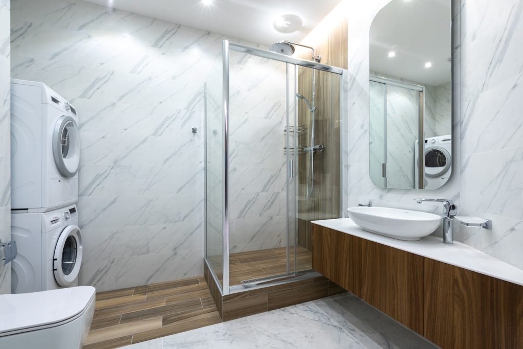 Renovación de baños y cocinas 2 Imagen de un cuarto de baño moderno, con paredes de mármol blanco con vetas grises, una elegante mampara de ducha de cristal y azulejos de color marrón a juego con el armario del lavabo.