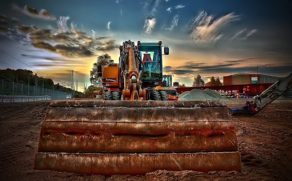Demolición y retirada de materiales antiguos 2 Imagen de una excavadora en primer plano con la pala de la excavadora destacada, mientras el cielo muestra una hermosa puesta de sol con tonos de grises y azules. La excavadora se encuentra en un terreno de tierra preparada para trabajar, y al fondo se aprecian montones de tierra y una nave industrial.